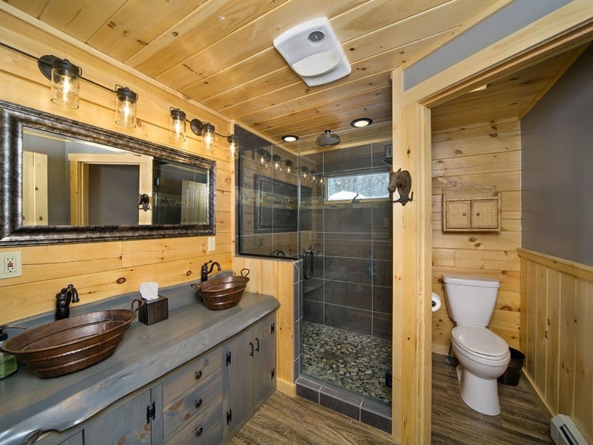 gray cabinet below brass bowl sinks in bathroom by tiled shower with glass door