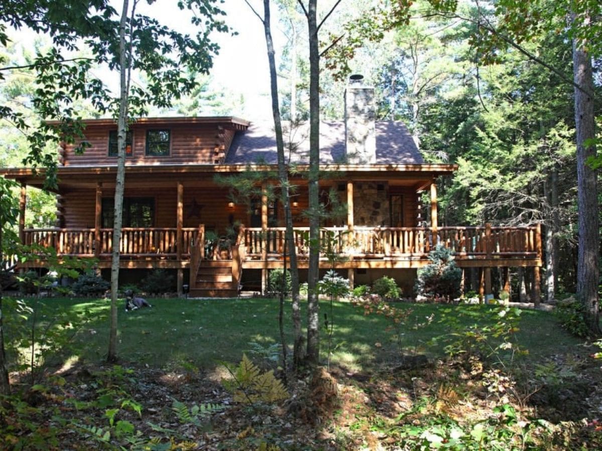 back of log cabin with large wrap around porch nestled in trees