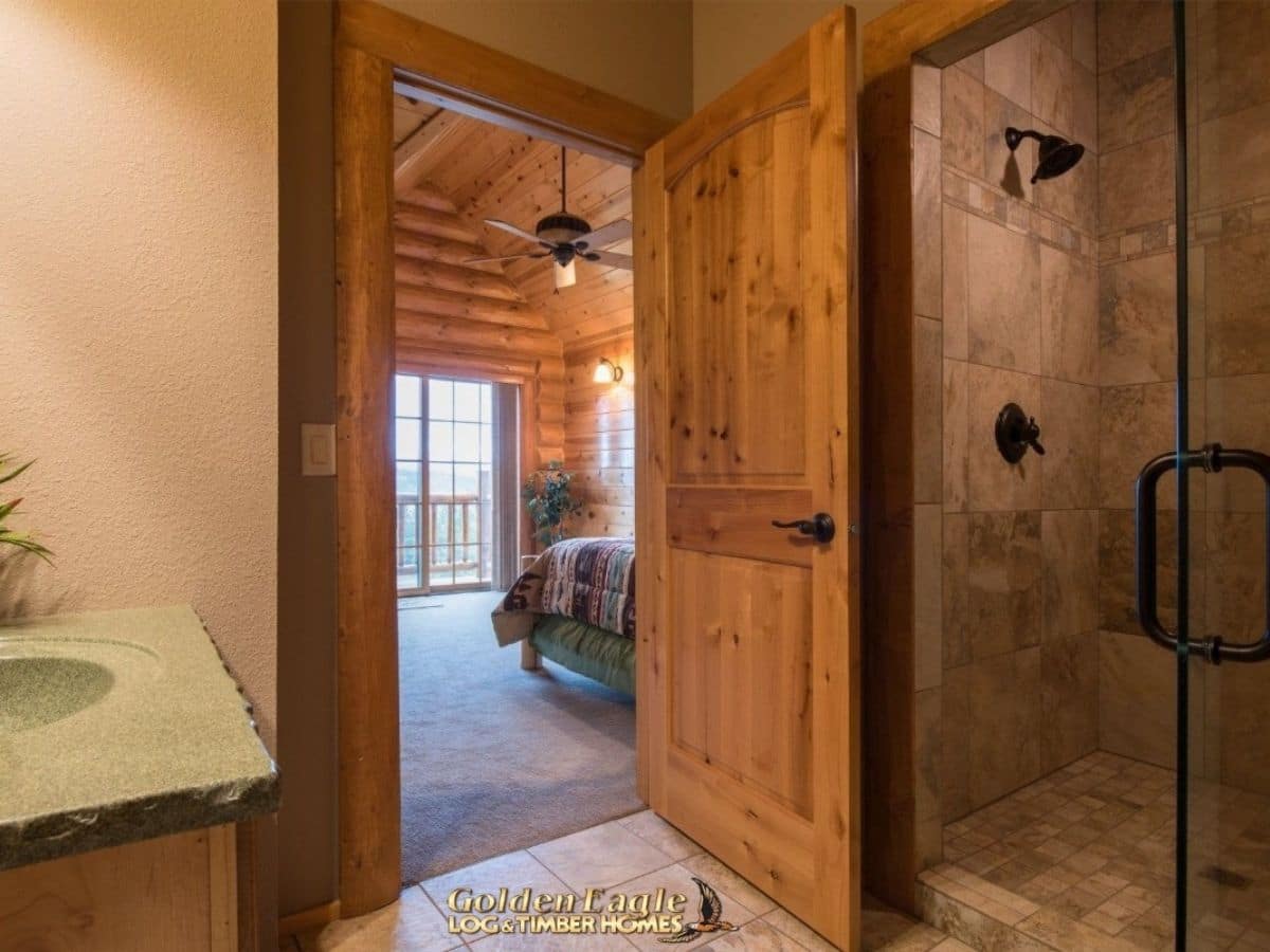 open door in bathroom with shower on right and view into bedroom with door to balcony