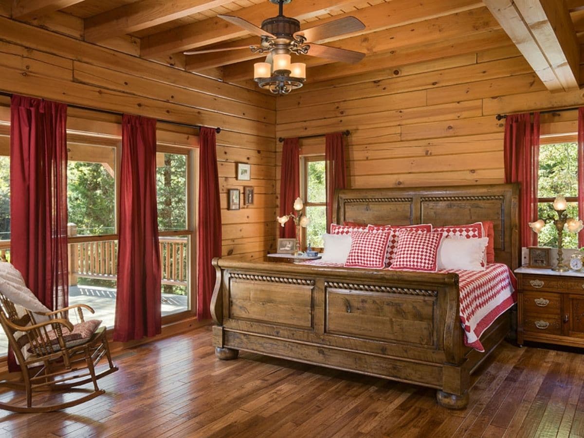 wood sleigh bed against log wall with red curtains on windows