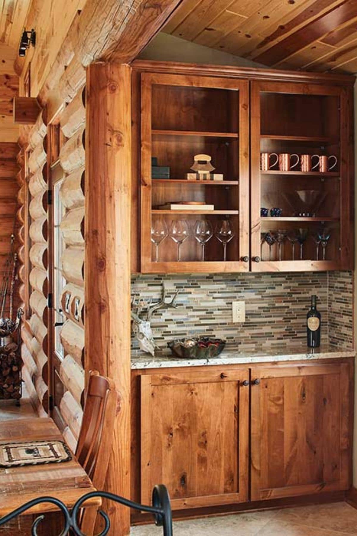 hutch with wine glasses against wall of log cabin with tile backsplash