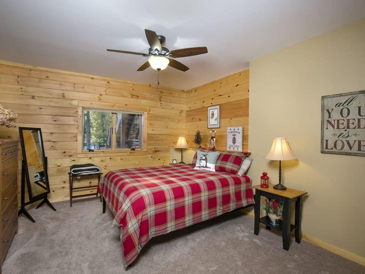 red plaid blanket on bed underneath white ceiling with ceiling fan and half log wall on left