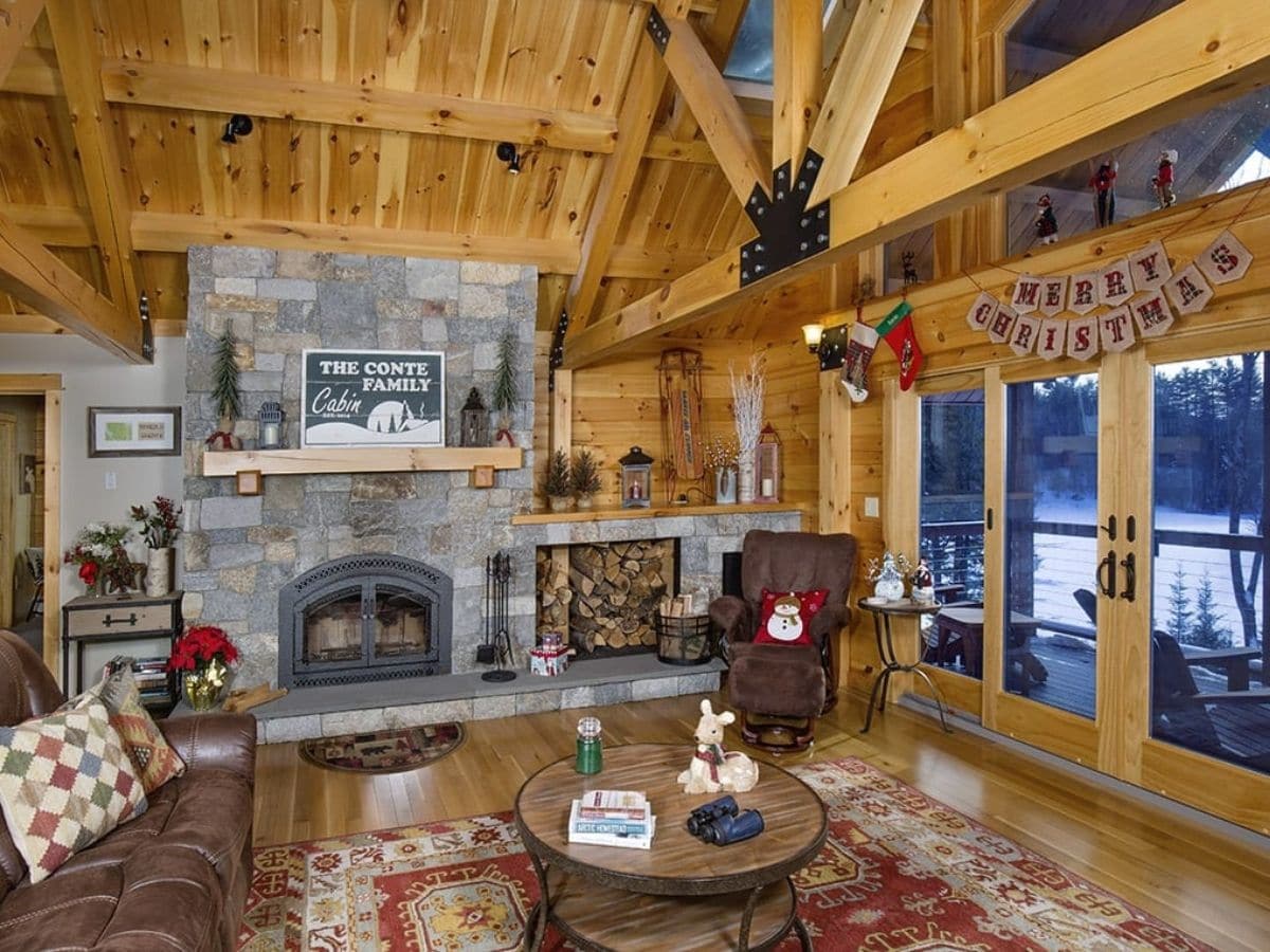 stone fireplace against wall of living room with exposed beam ceiling and picture windows to right of image