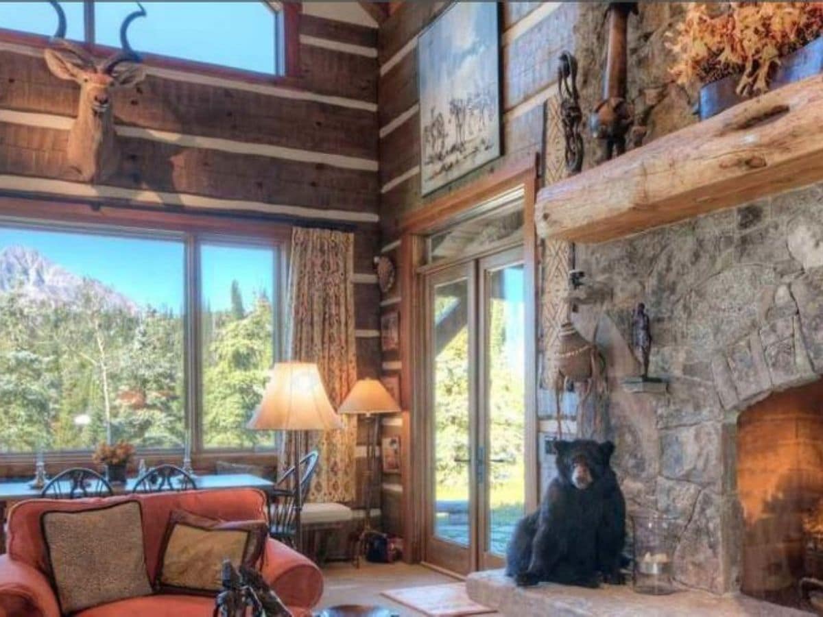 dark wood log cabin with picture windows against far wall with stone fireplace in foreground next to french doors