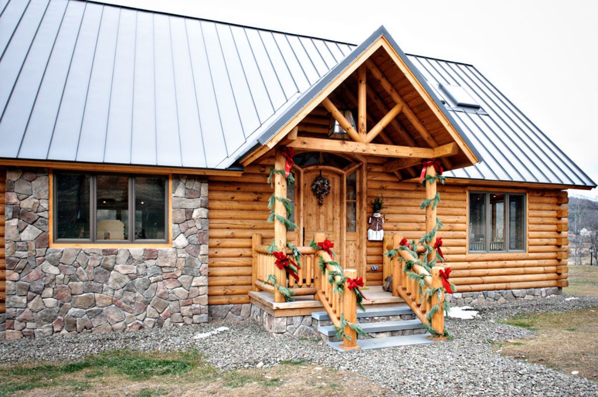 front of log cabin with partial stone wall on left and holiday decor on porch railings