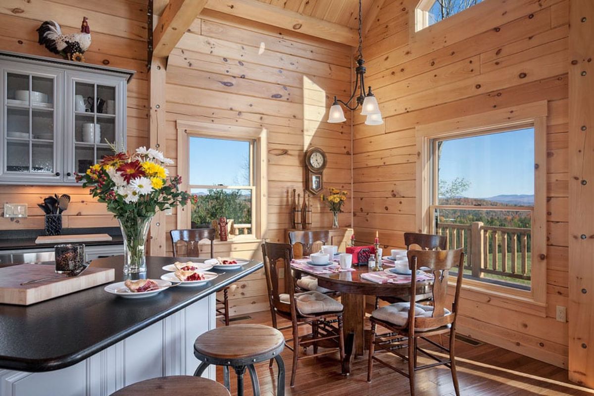 small dining table in corner of log cabin kitchen with stool in front next to island with black countertop