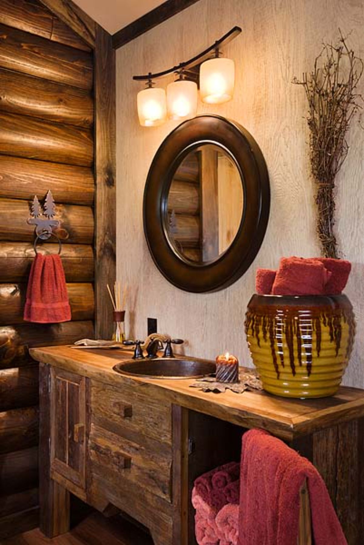 pottery on end of cabinet holding red towels with round mirror above sink against log walls