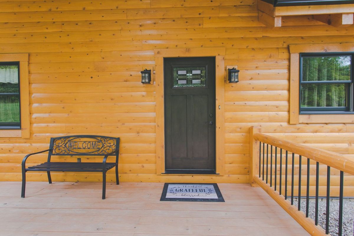 brown front door with welcome mat on front of log cabin with metal bench on left