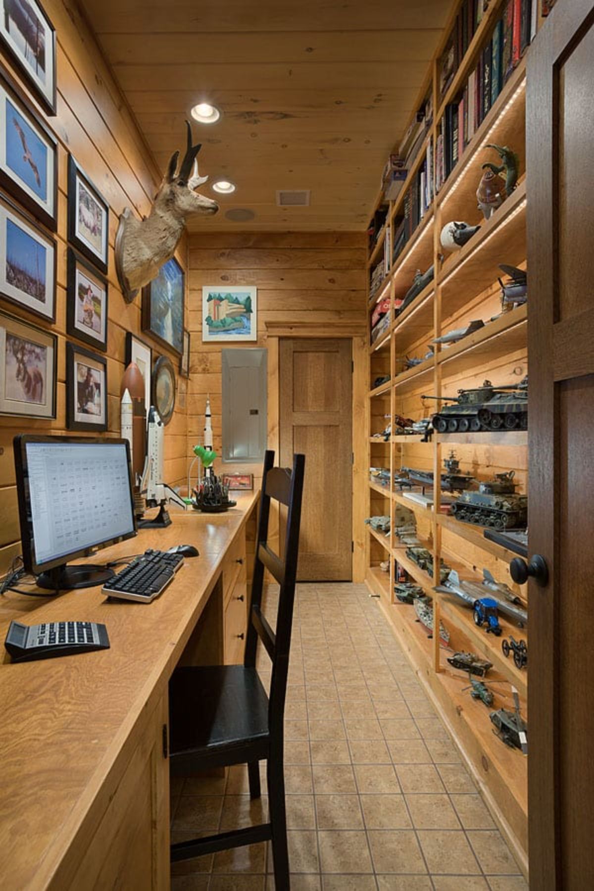 narrow room with shelves on one wall and pictures on the other with a desk lining the left side