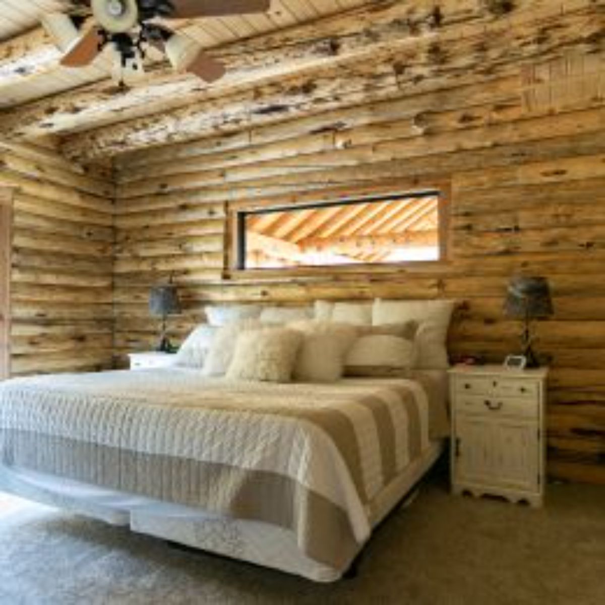 light bedding on bed against log cabin wall with long rectangular window above bed