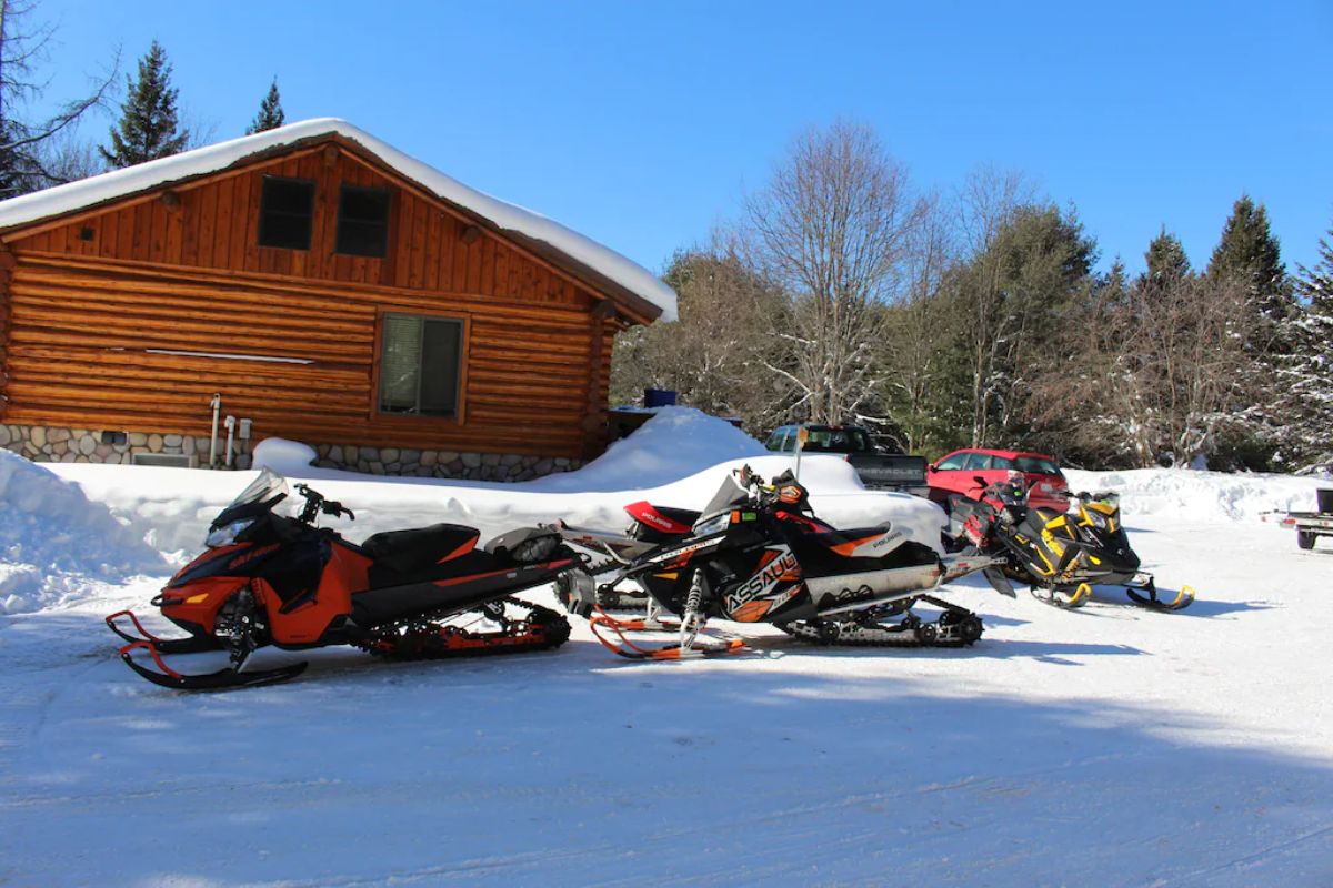 end of log cabin in snow with snowmobiles parked beside home