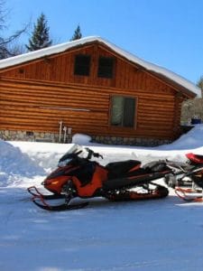 end of log cabin in snow with snowmobiles parked beside home