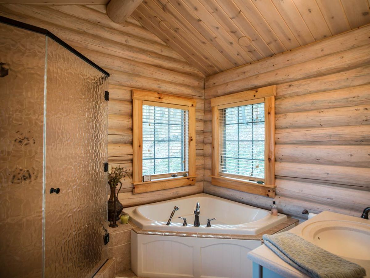 glass shower to right of white soaking tub in bathroom with log walls