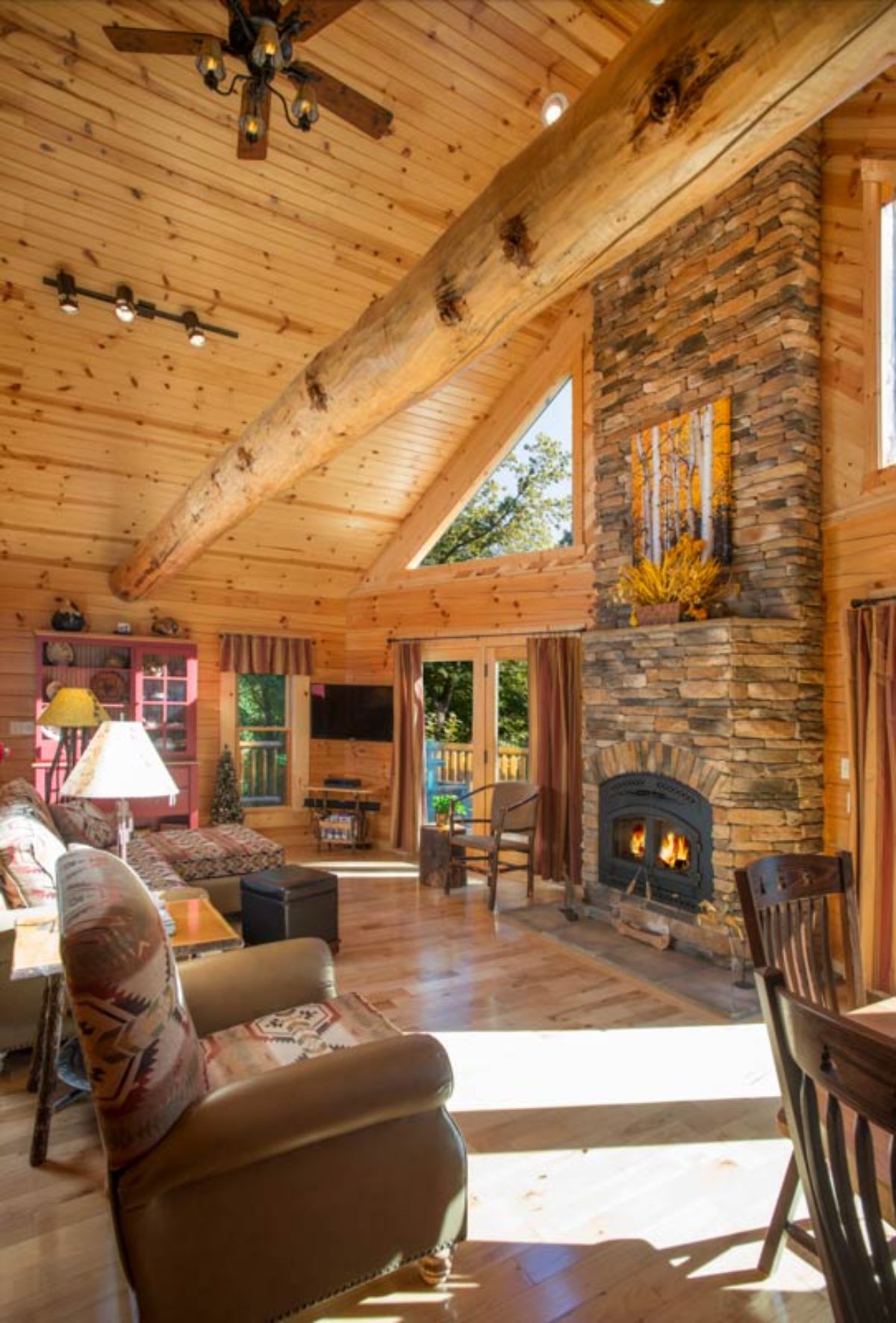 view into great room with stone fireplace between trapezoid windows and brown sofa across from fireplace