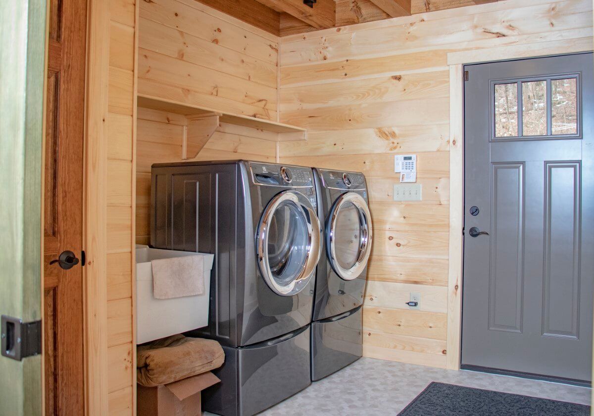 grey front load washer and dryer in corner against light wood log cabin walls with grey door to right