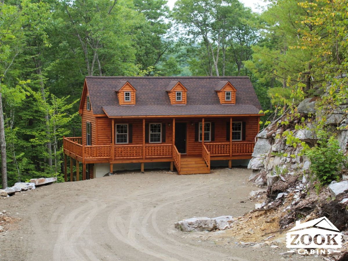 dark wood cabin with three dormer windows and shingle roof behind gravel driveway