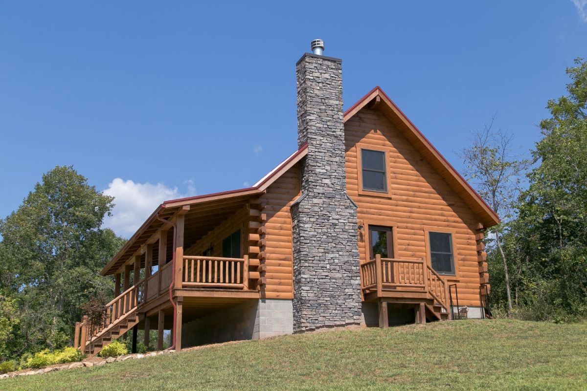 side of log cabin with stone chimney on the side by stairs to side door