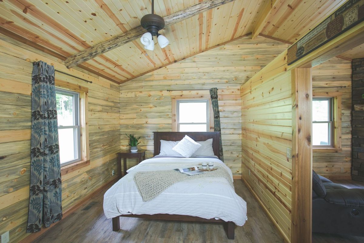 bed with dark wood frame and white linens against wall of windows in cabin bedroom