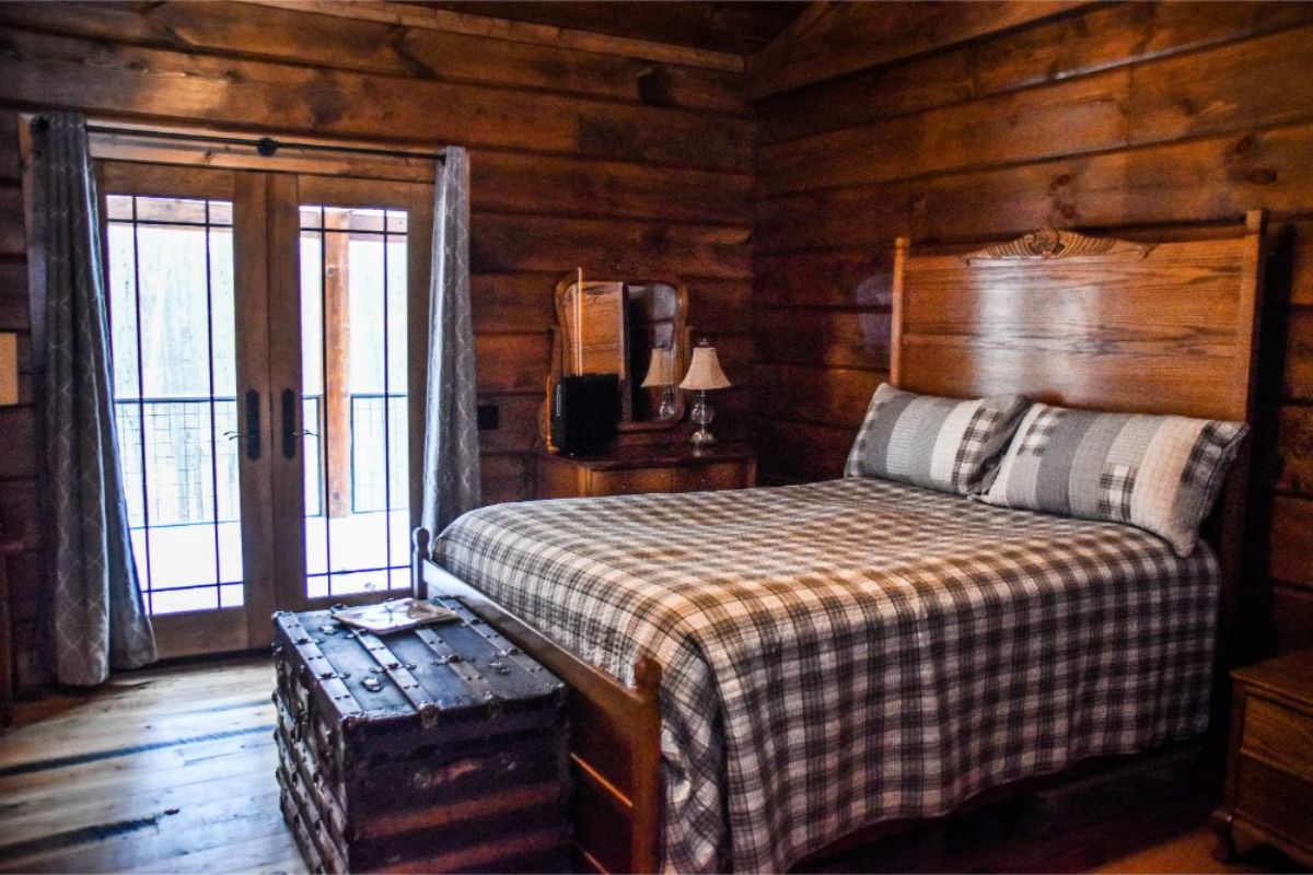 black and white bedding on bed in log cabin with trunk at end of bed and french doors on left