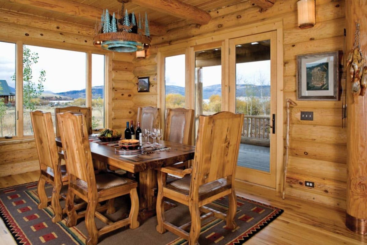 wood table and chairs under chandelier in log cabin with windows on three sides