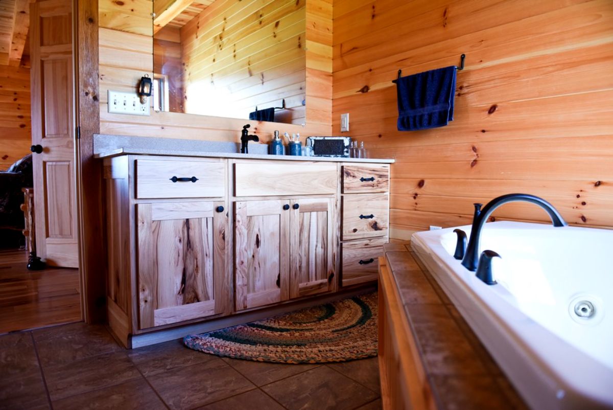white soaking tub on right of log cabin bathroom with light wood cabinet under mirror