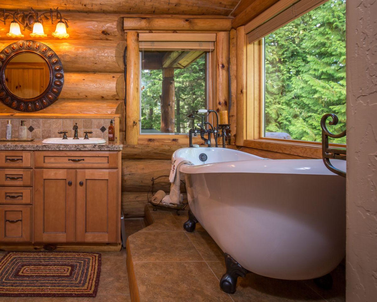 white soaking bathtub under picture window of log cabin with wood vanity to left of space