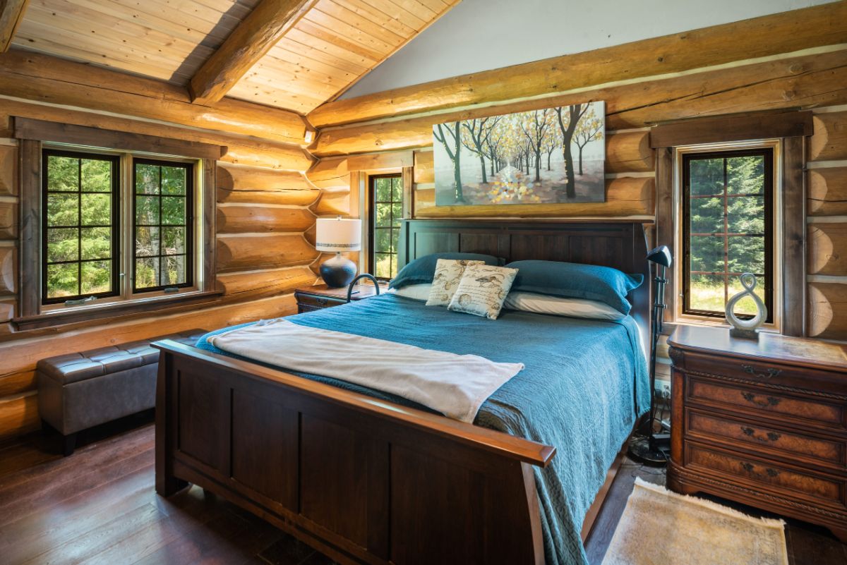 teal bedding on dark wood framed bed in log cabin bedroom with windows on both sides of the bed