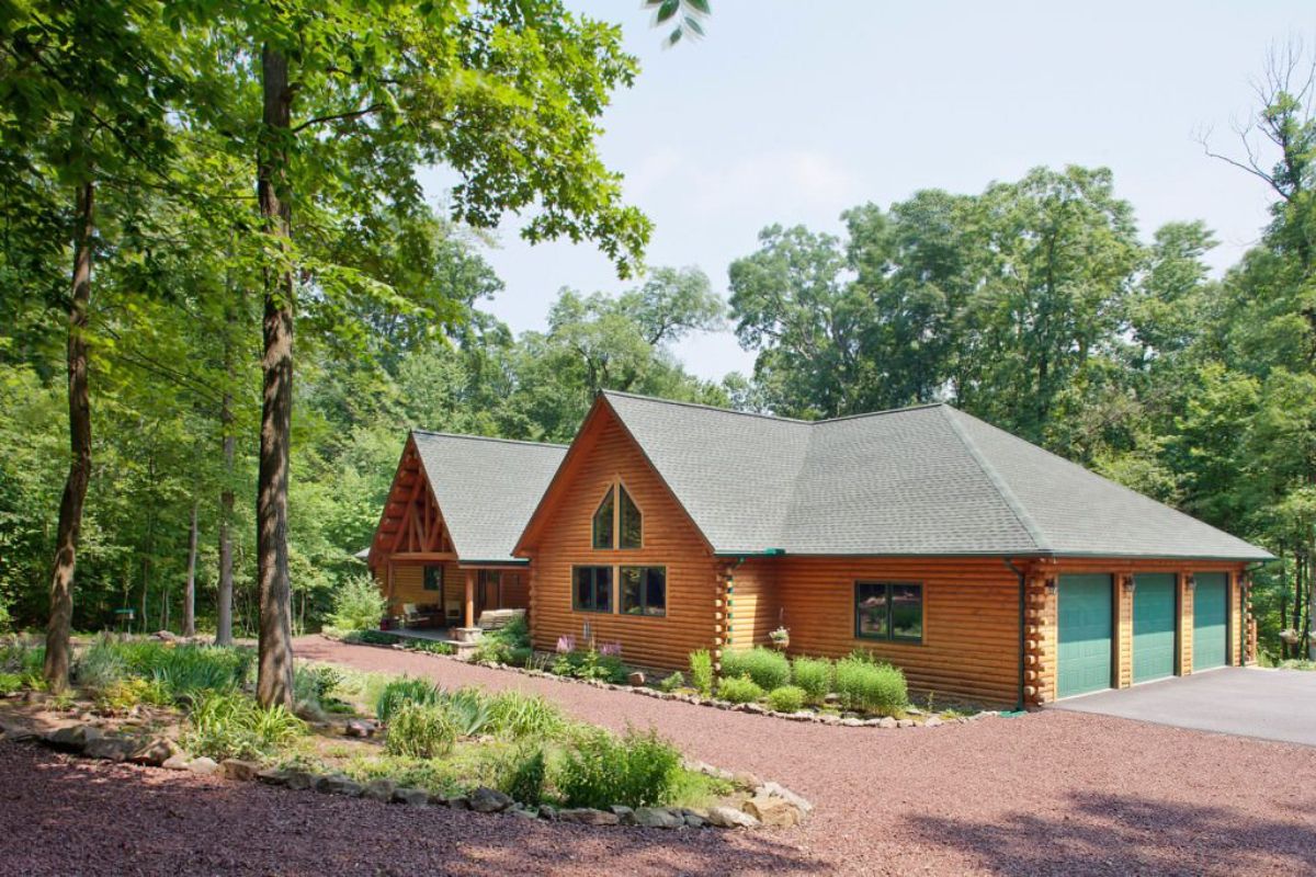 light wood cabin with green garage doors on right end behind brown rock driveway