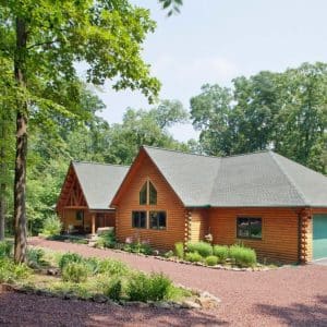 light wood cabin with green garage doors on right end behind brown rock driveway