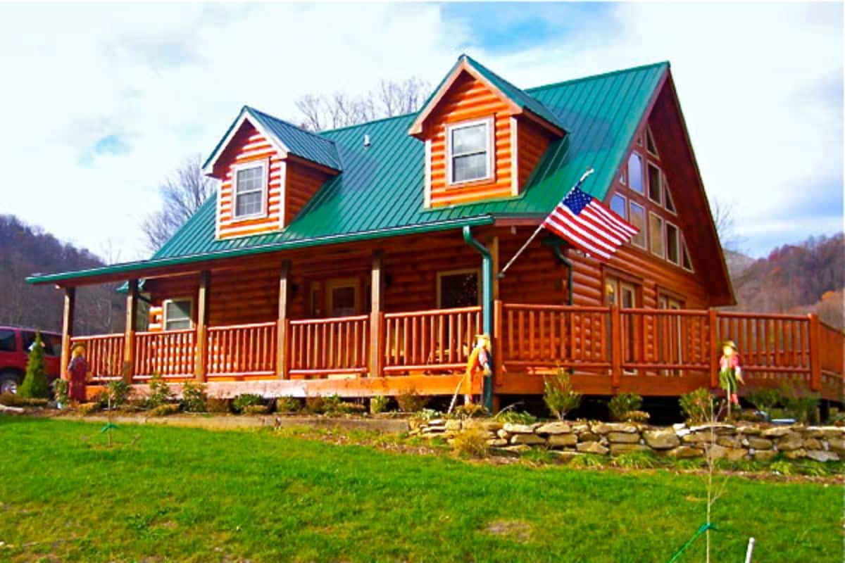 flag on end of porch in front of cabin with two dormer windows and green roof